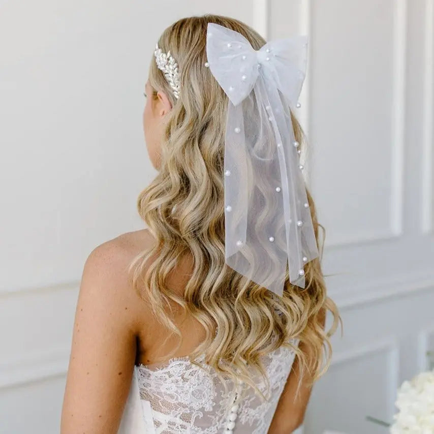 Back view of a woman with wavy blonde hair wearing an elegant tulle hair bow adorned with scattered pearls. The bow is delicate and airy, adding a touch of romance and sophistication to her bridal or special occasion hairstyle.