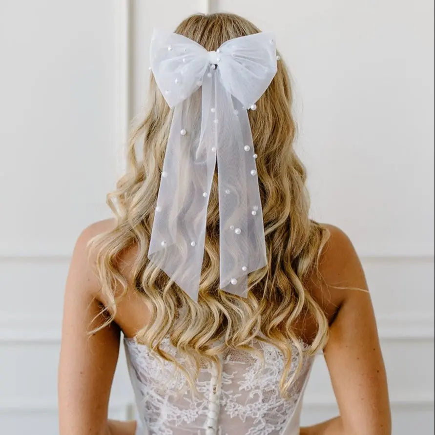 Back view of a woman with wavy blonde hair wearing an elegant tulle hair bow adorned with scattered pearls. The wedding veil bow is delicate and airy, adding a touch of romance and sophistication to her bridal or special occasion hairstyle.